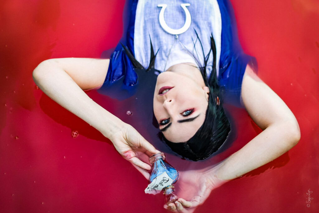 Cosplayer als Alice aus dem Spiel Alice Madness Returns liegt in einem rot gefärbten Wasser und hält eine "Trink Mich" Flasche in den Händen.